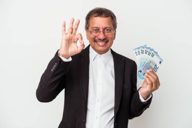 Middle aged indian business man holding bills isolated on white background cheerful and confident showing ok gesture.