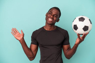 Mavi arka planda izole bir şekilde futbol oynayan genç Afrikalı Amerikalı bir adam bir avuç içinde fotokopi alanı ve belinde başka bir el tutuyor..