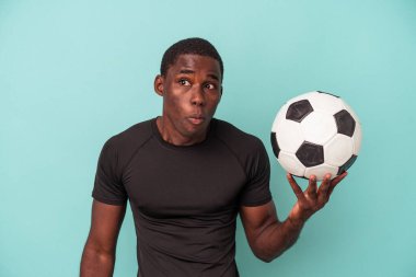 Mavi arka planda izole bir şekilde futbol oynayan genç Afrikalı Amerikalı omuz silker ve gözleri açık kalır..