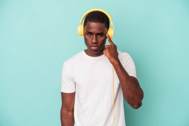 Genç Afrikalı Amerikalı adam mavi arka planda izole edilmiş müzik dinliyor. Şakağı parmağıyla işaret ediyor, düşünüyor, bir göreve odaklanıyor..