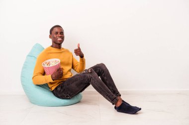 Genç Afrikalı Amerikalı bir adam beyaz arka planda izole edilmiş patlamış mısır yiyor gülümsüyor ve başparmağını kaldırıyor.