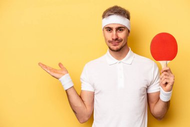 Young caucasian man holding ping pong rackets isolated on yellow background showing a copy space on a palm and holding another hand on waist.