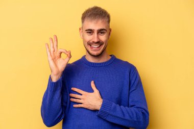 Sarı arka planda izole edilmiş genç beyaz bir adam bir göz kırptı ve elleriyle iyi bir jest yaptı..