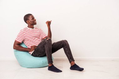 Genç Afrikalı Amerikalı bir adam beyaz arka planda izole edilmiş bir şekilde oturuyor. Baş parmak uzakta, gülüyor ve kaygısız..