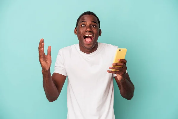 一个年轻的非裔美国人拿着一部蓝色背景的手机 带着惊喜 兴奋地举起双手 — 图库照片