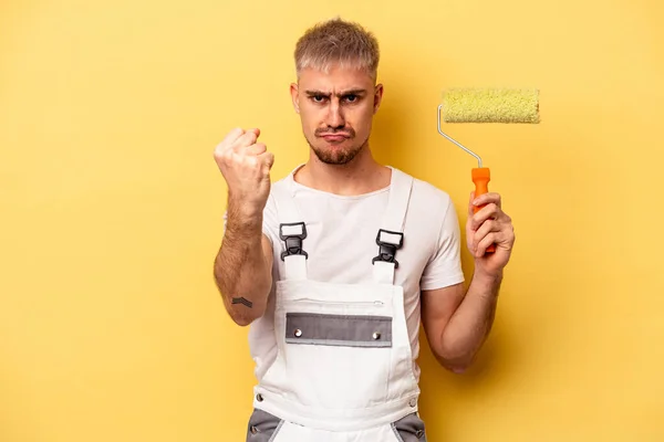 Young painter man isolated on yellow background showing fist to camera, aggressive facial expression.