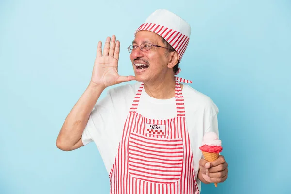 Senior american ice cream man holding an ice cream isolated on blue background shouting and holding palm near opened mouth.