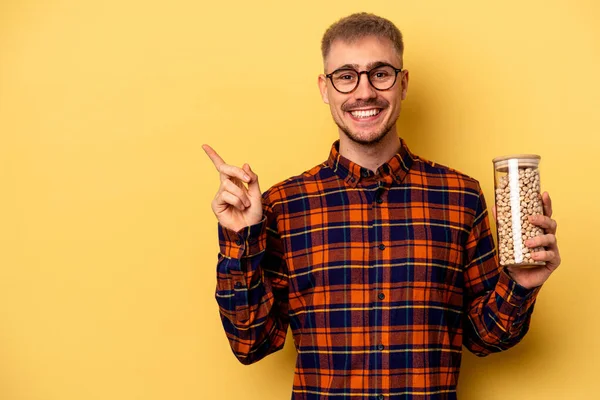 Young Caucasian Man Holding Chickpeas Isolated Yellow Background Smiling Pointing — Zdjęcie stockowe