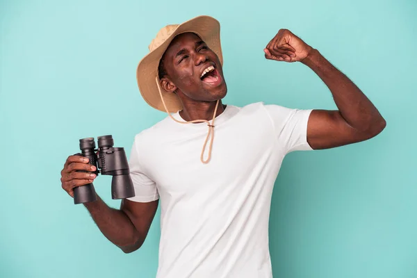 年轻的非洲裔美国男子手持双筒望远镜 在胜利后举着蓝色背景的拳头 赢得了胜利 — 图库照片