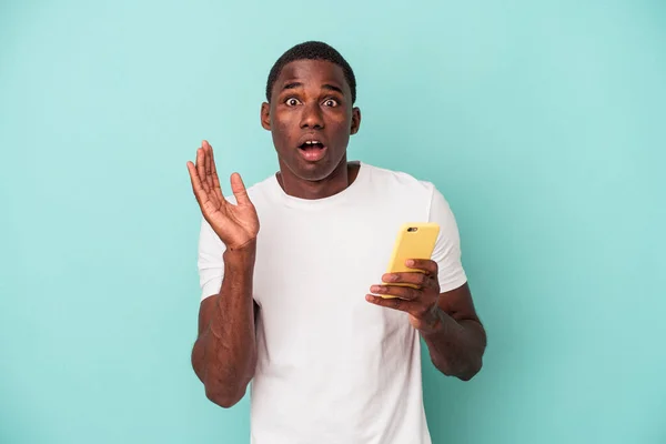 拿着一部蓝色背景的手机的年轻的非裔美国人感到惊讶和震惊 — 图库照片