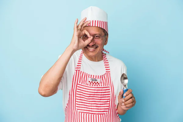 Senior indian ice cream man holding a scoop isolated on blue background excited keeping ok gesture on eye.