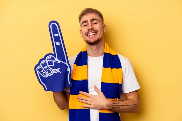 Young sports fan caucasian man isolated on yellow background laughing and having fun.