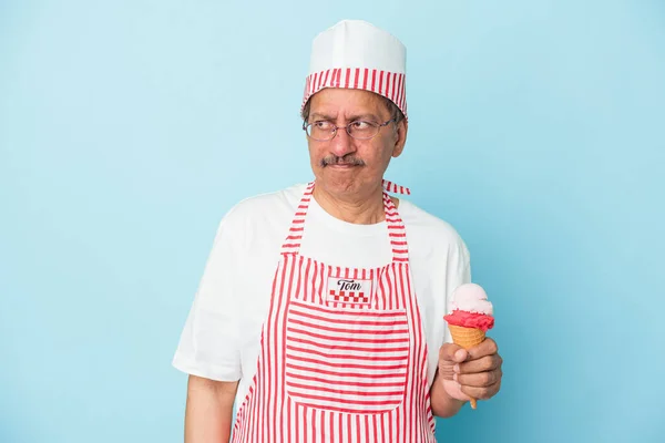 Senior american ice cream man holding an ice cream isolated on blue background confused, feels doubtful and unsure.