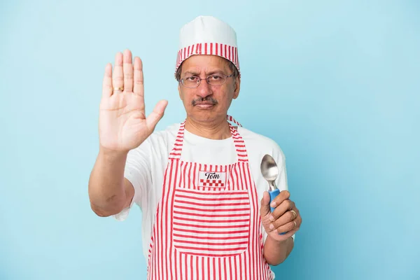 Senior indian ice cream man holding a scoop isolated on blue background standing with outstretched hand showing stop sign, preventing you.