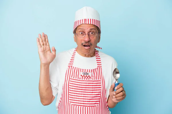 Senior indian ice cream man holding a scoop isolated on blue background surprised and shocked.