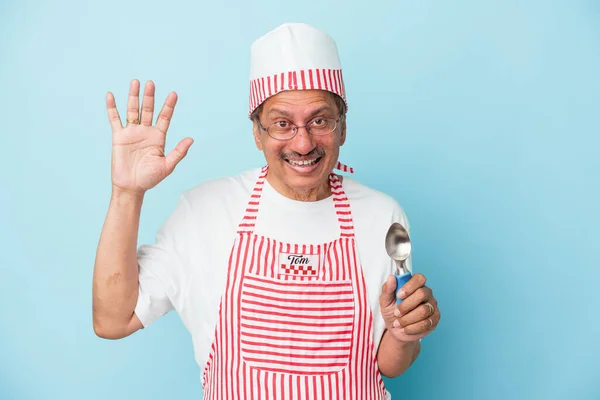 Senior indian ice cream man holding a scoop isolated on blue background receiving a pleasant surprise, excited and raising hands.