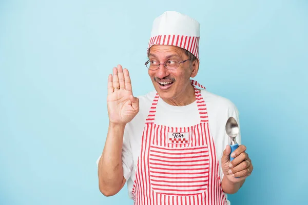 Senior indian ice cream man holding a scoop isolated on blue background is saying a secret hot braking news and looking aside