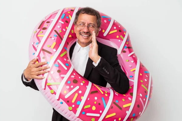 Senior indian business man holding inflatable donut isolated on white background is saying a secret hot braking news and looking aside