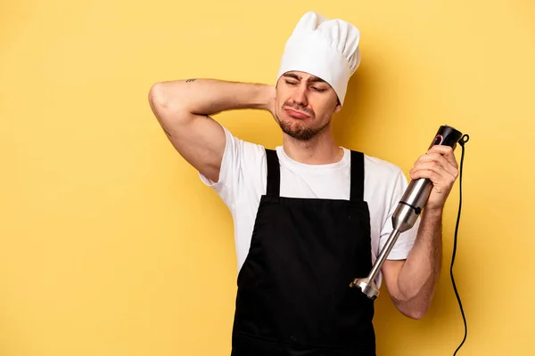 Young caucasian chef man holding blender isolated on yellow background touching back of head, thinking and making a choice.