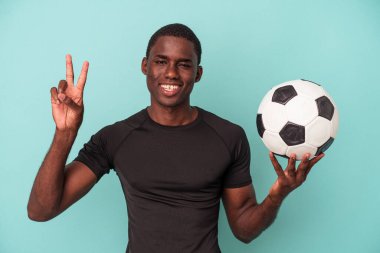 Mavi arka planda izole bir şekilde futbol oynayan genç Afrikalı Amerikalı iki numarayı parmaklarıyla gösteriyor..