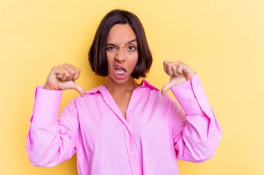 Sarı arka planda izole edilmiş genç melez kadın baş parmağını aşağı gösteriyor ve hoşlanmadığını ifade ediyor..