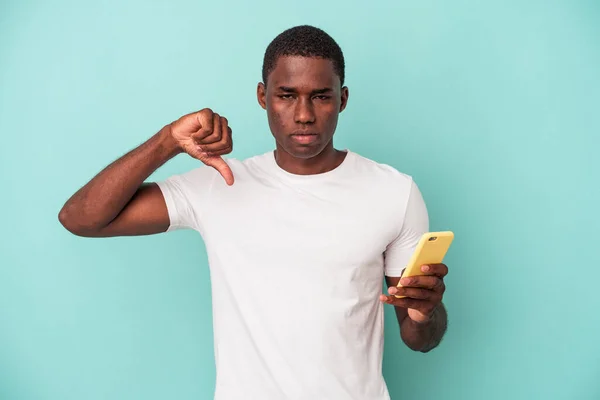 年轻的非裔美国人拿着一部蓝色背景的手机 表现出不喜欢的姿态 大拇指垂下 不同意的概念 — 图库照片
