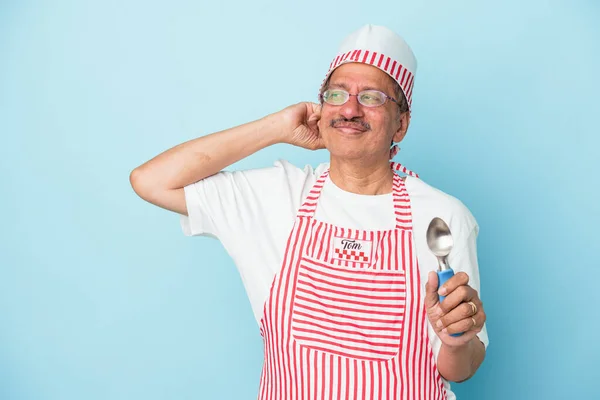 Senior indian ice cream man holding a scoop isolated on blue background touching back of head, thinking and making a choice.