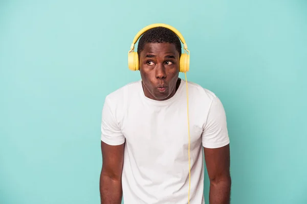 若いアフリカ系アメリカ人男性音楽を聞いて隔離された青の背景Shrugs肩とオープン目混乱 — ストック写真