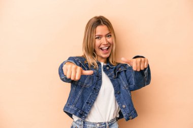 Beyaz tenli genç bir kadın bej arka planda izole edilmiş. Baş parmağını kaldırıyor, gülümsüyor ve kendinden emin..