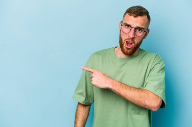 Mavi arka planda izole edilmiş genç beyaz adam yan tarafı işaret ediyor.