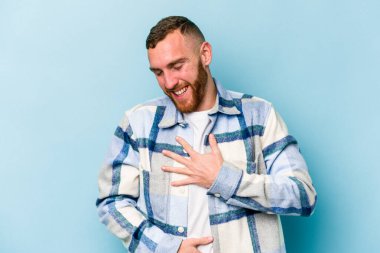 Mavi arka planda izole edilmiş genç beyaz adam mutlu bir şekilde gülüyor ve ellerini karnının üstünde tutarak eğleniyor..