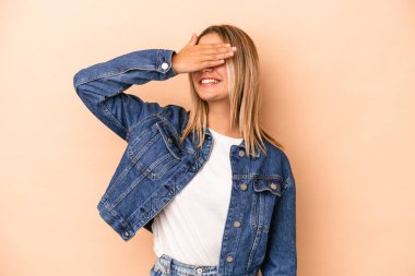 Beyaz tenli, bej arka planda izole edilmiş genç bir kadın elleriyle gözlerini kapatıyor, gülümseyerek bir sürpriz bekliyor..