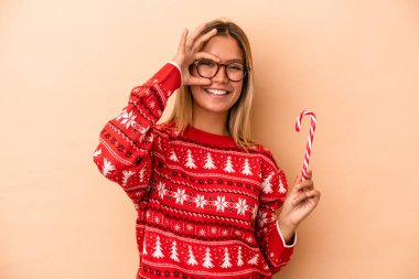 Young caucasian woman holding a christmas stick isolated on beige background excited keeping ok gesture on eye.