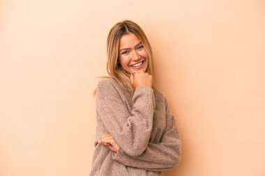 Beyaz tenli genç bir kadın bej arka planda izole edilmiş mutlu ve kendinden emin gülümsüyor, elleriyle çenesine dokunuyor..