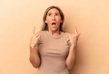 Beyaz tenli genç bir kadın, bej arka planda izole edilmiş, ağzı açık, üst tarafı işaret ediyor..
