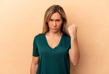 Beyazlı genç bir kadın bej arka planda izole edilmiş, yumruğunu kameraya gösteriyor, agresif yüz ifadesi var..