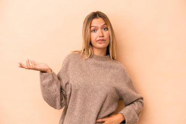Beyaz tenli genç bir kadın bej arka planda izole edilmiş sorgulama sırasında omuz silkiyor ve şüphe duyuyor..