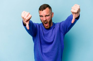 Mavi arka planda izole edilmiş genç beyaz bir adam baş parmağını aşağı gösteriyor ve hoşlanmadığını ifade ediyor..