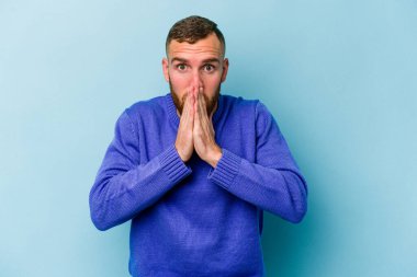 Kafkasyalı genç adam mavi arka planda izole edilmiş ağzı kapalı ve elleri endişeli görünüyor..