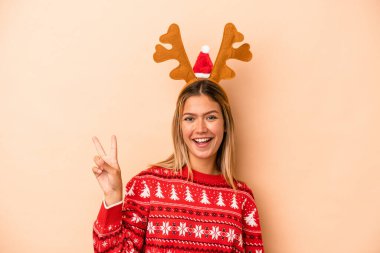 Young caucasian woman wearing a christmas reindeer hat isolated on beige background joyful and carefree showing a peace symbol with fingers.