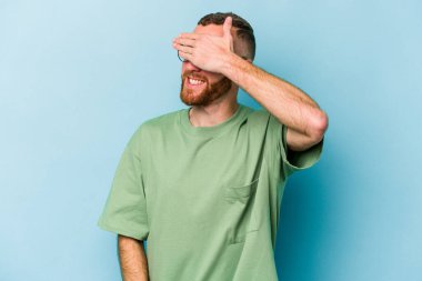 Mavi arka planda soyutlanmış genç beyaz bir adam elleriyle gözlerini kapatıyor, geniş bir gülümsemeyle bir sürpriz bekliyor..