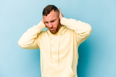 Mavi arka planda izole edilmiş genç beyaz adam kulaklarını elleriyle kapatıyor..