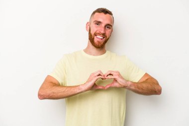 Beyaz arka planda izole edilmiş genç beyaz bir adam gülümsüyor ve elleriyle kalp şekli gösteriyor..