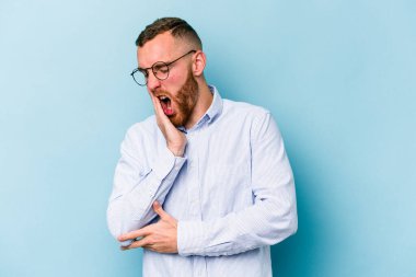 Mavi arka planda izole edilmiş genç beyaz adam esniyor. Ağzı eliyle kapatan yorgun bir hareket sergiliyor..