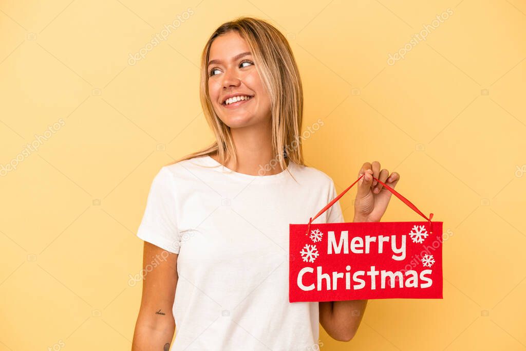 Young caucasian woman holding a merry christmas placard isolated on yellow background looks aside smiling, cheerful and pleasant.