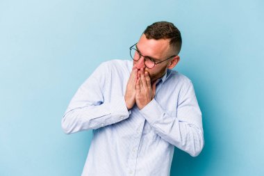 Mavi arka planda izole edilmiş genç beyaz bir adam bir şeye gülüyor, ağızlarını ellerle kapatıyor..