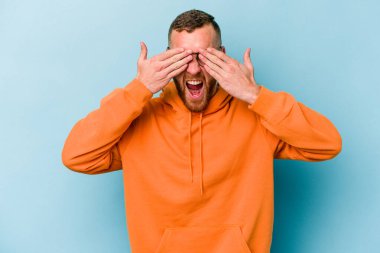 Mavi arka planda soyutlanmış genç beyaz bir adam elleriyle gözlerini kapatıyor, geniş bir gülümsemeyle bir sürpriz bekliyor..