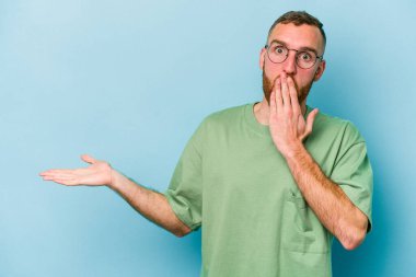 Mavi arka planda izole edilmiş genç beyaz adam, avuç içi fotokopi alanını elinde tutmaktan etkilenmiş..