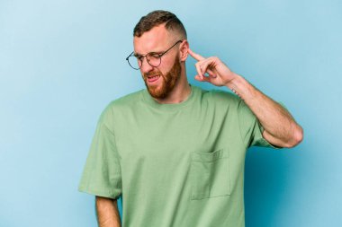 Genç beyaz adam mavi arka planda izole edilmiş parmaklarıyla kulaklarını kapatıyor, gürültülü bir ortam tarafından stresli ve umutsuz..