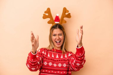 Young caucasian woman wearing a christmas reindeer hat isolated on beige background receiving a pleasant surprise, excited and raising hands.